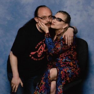 Against a blue backdrop, Richmond and Carrie Fisher have their arms around each other. Richmond wears a black t-shirt and trousers, with rectangular framed glasses, while Carrie wears a colourful, sparkly red and blue dress. She is also wearing rectangular glasses, and they both have their hair in ponytails.