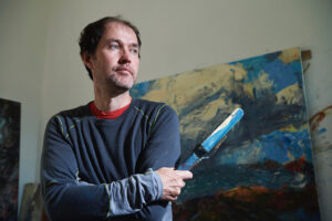 Allan is a white man with short brown hair. He is standing, looking away to the right with his arms folded. In one hand, he holds a paint sponge/brush covered in blue paint and a palette knife. He's wearing a navy long sleeved top with a red top peeking out at his neck underneath. A large painted canvas sits behind him made up of yellows, blues and browns.