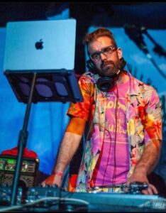 Ross' headshot. He is DJ'ing using decks and a laptop on a stand. He is a white man with brown hair and a beard, wearing glasses and a bright pink, orange and yellow Hawaiian style shirt, with a pink top underneath. Ross has headphones around his neck.