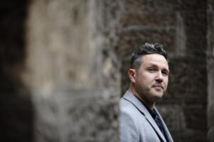 Alan's headshot is taken against a tall, grey brick wall. He is a white man with black hair, and he is wearing a grey suit jacket and a dark coloured shirt. He can be seen from the chest up. Alan looks thoughtfully into the distance towards the camera.