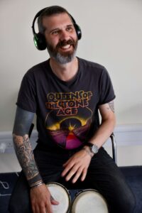 Dave's headshot. He is sitting with small drums between his knees as he smiles widely up at something beyond the camera. Dave is wearing all black, with a 'Queens of the Stone Age' top on, and wireless headphones over his ears.