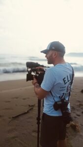 Graeme's headshot. Graeme is standing on a beach holding a film camera towards the water, with a photography camera slung on a strap over his shoulder. He is wearing a navy cap, light blue t-shirt with text on the back and shorts.