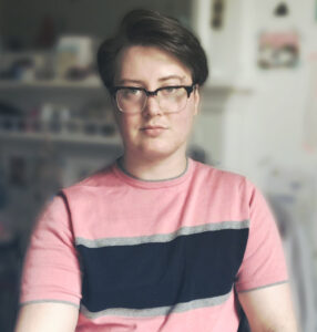 Jem's headshot. They are wearing a pink t-shirt with a thick navy blue stripe across the front, sandwiched by two slimmer grey lines. Jem has short brown hair swept over their face, and thick square glasses.