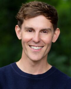 A front-facing photograph of Joseph Elliott. He is a white man with brown eyes and brown hair swept across his forehead. He smiles widely, and is wearing a round-neck navy blue t-shirt.