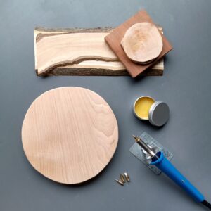 a selection of carved wood on a table, with a wood burning pen and pyrography materials arranged aesthetically on a grey backdrop