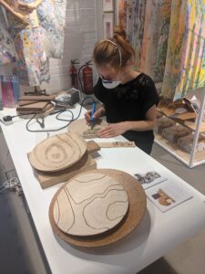 Lee at work in a studio. She wears a dust mask and has her hair tied back while she works on a piece of wood. 2 other round pieces of art sit away from her on the white table towards the camera.