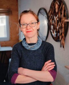 Lee's headshot. Lee is a white woman with light hair tied back off her face. She wears blue square rimmed glasses and has a friendly smile. Her arms are crossed, and she is wearing a navy jumper with purple cuffs rolled up by her elbows, with a sky blue patterned buff around her neck.