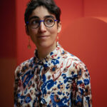 A person with short brown hair, round glasses and wearing a white, blue, red and yellow patterned top standing in front of red background.