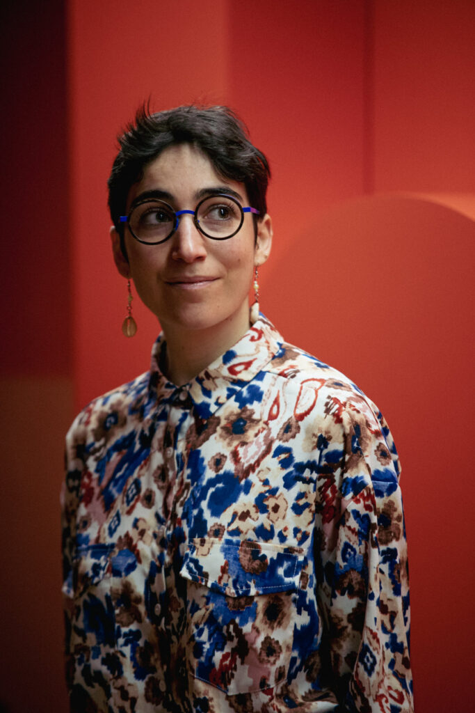 A person with short brown hair, round glasses and wearing a white, blue, red and yellow patterned top standing in front of red background.