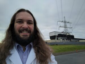 A selfie in front of a powerplant. John is a white man with shoulder length brown hair and a beard. He has a wide, friendly smile and wears a pale blue shirt and cream blazer. The sky is grey and cloudy.